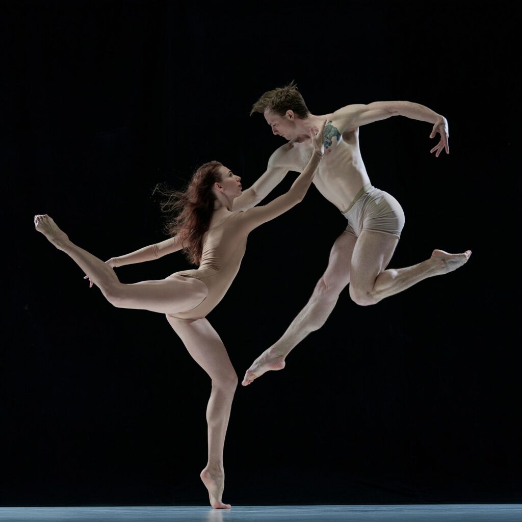 Two Columbia Classical Ballet dancers in nude costumes mid-leap against a black background, Project 27